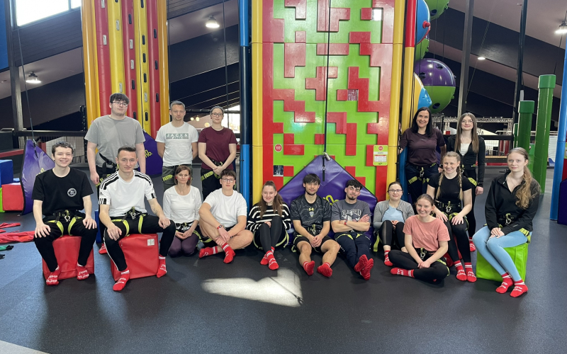 Gruppenfoto der Auszubildenden in Klettergurten und roten Socken vor bunten Kletterw&auml;nden im Jump & Climb in Hof