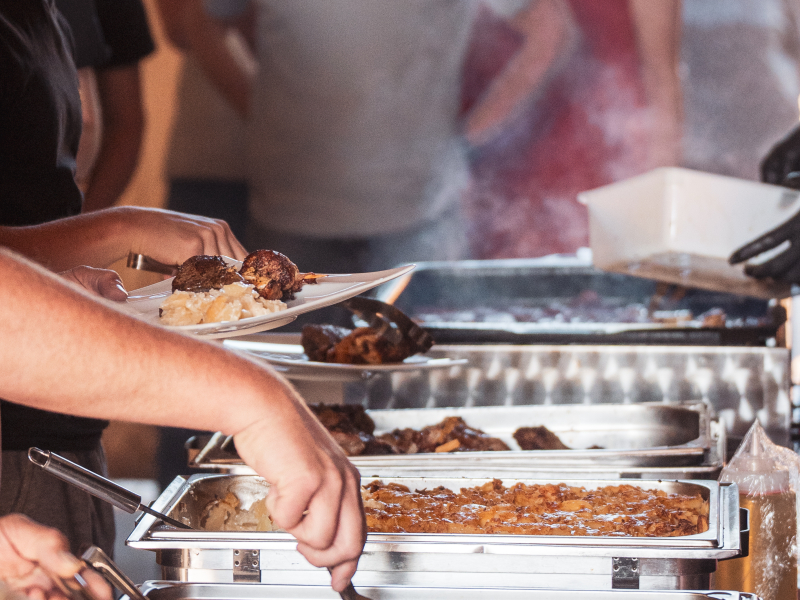 Grillbuffet mit Fleisch und Gem&uuml;se in Warmhalteschalen, dahinter stehen Kolleginnen und Kollegen an
