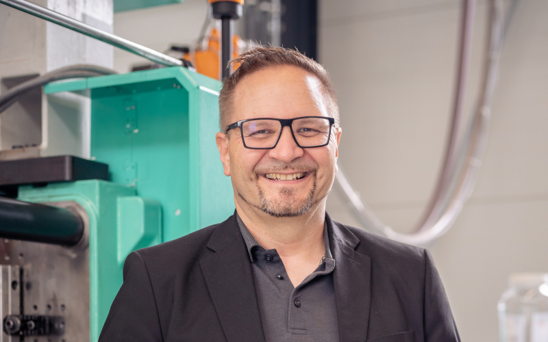 Portr&auml;t von Martin Rei&szlig; mit einem Kunststoffteil in der Hand vor einer Spritzgussmaschine